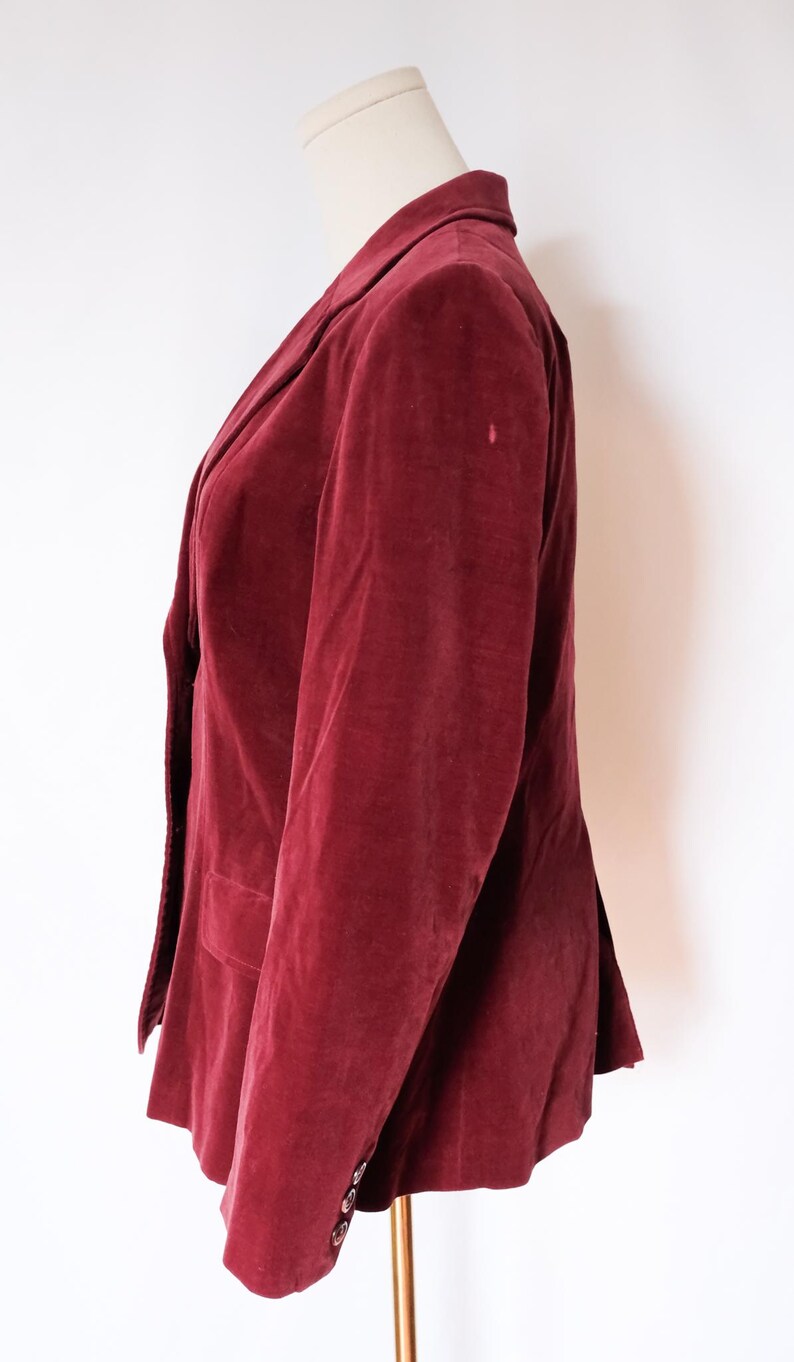 May include: A burgundy velvet blazer displayed on a mannequin. The blazer features a notched lapel, a two-button closure, and a single chest pocket. The sleeves have three buttons at the cuff. The jacket is a rich, deep red color.