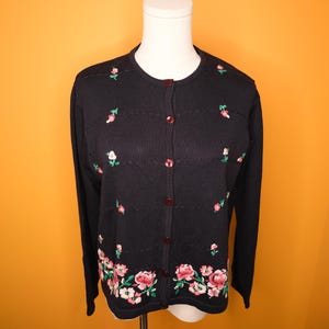 May include: A black cardigan sweater with a floral embroidery design. The sweater has a button-up front with small red buttons and long sleeves. The floral embroidery features pink and white flowers with green leaves.