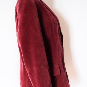 May include: A burgundy velvet blazer displayed on a mannequin. The blazer has a classic notched lapel, a two-button closure, and a single pocket. The fabric has a soft, textured appearance.