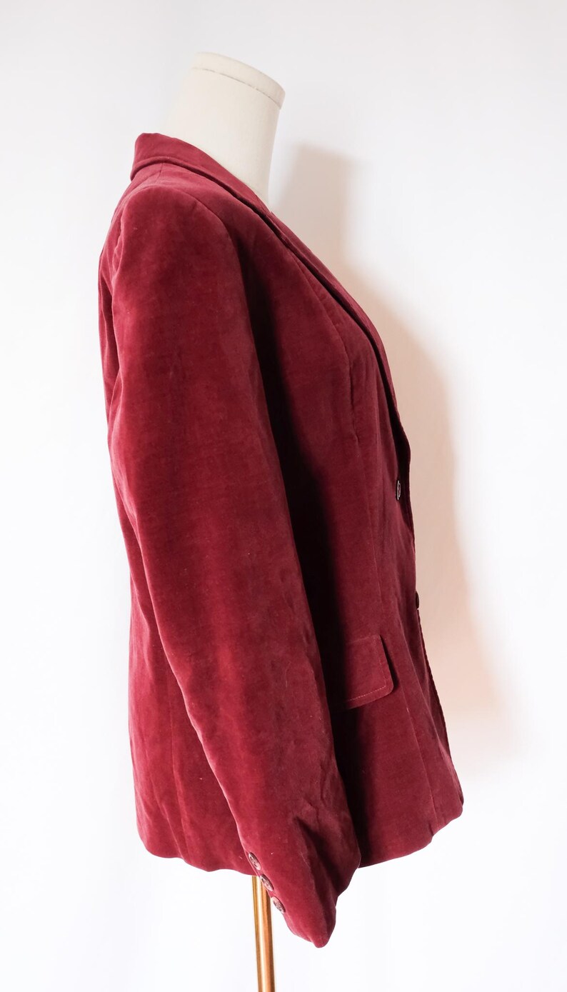 May include: A burgundy velvet blazer displayed on a mannequin. The blazer has a classic notched lapel, a two-button closure, and a single pocket. The fabric has a soft, textured appearance.
