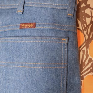 May include: Close-up of light blue denim jeans with the brand "Wrangler" visible on a small brown tag. The jeans have classic stitching details in a contrasting tan color. The image shows the back pocket and waistband.