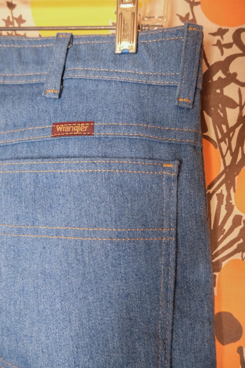 May include: Close-up of light blue denim jeans with the brand "Wrangler" visible on a small brown tag. The jeans have classic stitching details in a contrasting tan color. The image shows the back pocket and waistband.