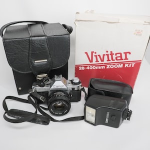 May include: A vintage Canon AE-1 camera kit with a black leather case, flash, and a Vivitar 28-400mm zoom kit box. The camera is silver and black with a lens attached. The kit includes a black camera strap.
