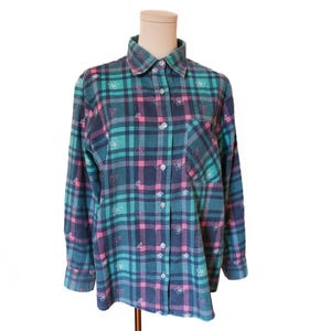 May include: A long-sleeved plaid shirt in teal, pink, and navy blue, adorned with small white floral embroidery. The shirt features a collared neckline, a single chest pocket, and button-down closure. The fabric has a soft, slightly worn appearance.