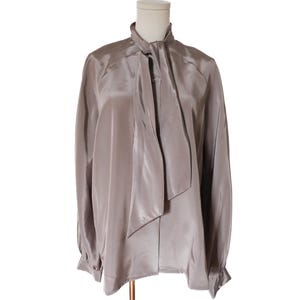 May include: A long-sleeved, taupe-colored blouse with a necktie detail. The blouse is made of a shiny, silky material and has a button closure at the neckline. The sleeves have buttoned cuffs.