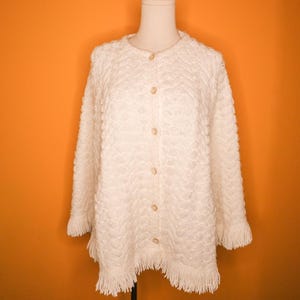 May include: Cream-colored, button-down cardigan with a textured, patterned design. The long-sleeved sweater features fringe at the hem and cuffs. The garment is displayed on a mannequin against an orange background.