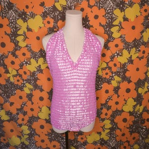 May include: A pink halter top with a textured, geometric pattern. The top has a deep V-neck and is displayed on a mannequin. The background features a floral pattern with orange and yellow flowers.