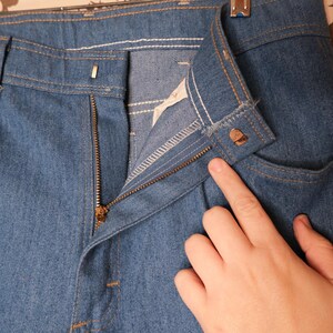 May include: Close-up of blue denim pants with a zipper and button closure. The pants have a classic five-pocket design with visible stitching in a contrasting color. The zipper is partially open, and the button is fastened.