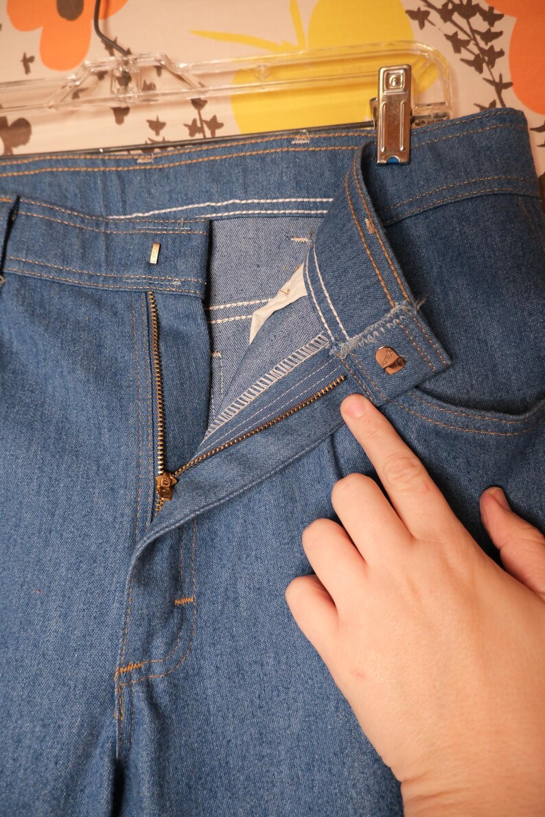 May include: Close-up of blue denim pants with a zipper and button closure. The pants have a classic five-pocket design with visible stitching in a contrasting color. The zipper is partially open, and the button is fastened.