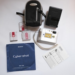 May include: A silver Sony Cyber-shot DSC-T100 digital camera with a wrist strap, a black camera case, a battery charger, batteries, and a manual. The camera has a 5x zoom lens and 1080 resolution. The camera case is black with a silver zipper.