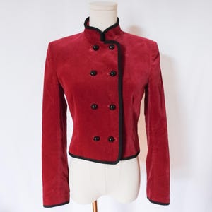 May include: A red velvet jacket with a mandarin collar and double-breasted button closure. The jacket features black trim along the collar, front, and hem. The jacket is displayed on a mannequin.