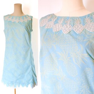 May include: A sleeveless, light blue dress with a pineapple pattern. The dress features a white lace trim around the neckline and hem. The neckline is adorned with embroidered pineapple shapes.