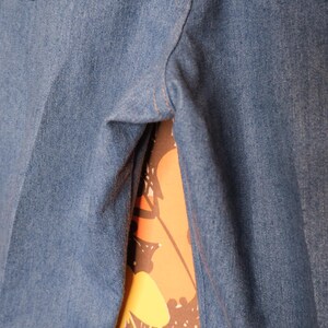 May include: Close-up of blue denim pants with two pockets and tan stitching. The pants are made of a medium-weight denim fabric. The background shows a floral pattern in orange, yellow, and brown.