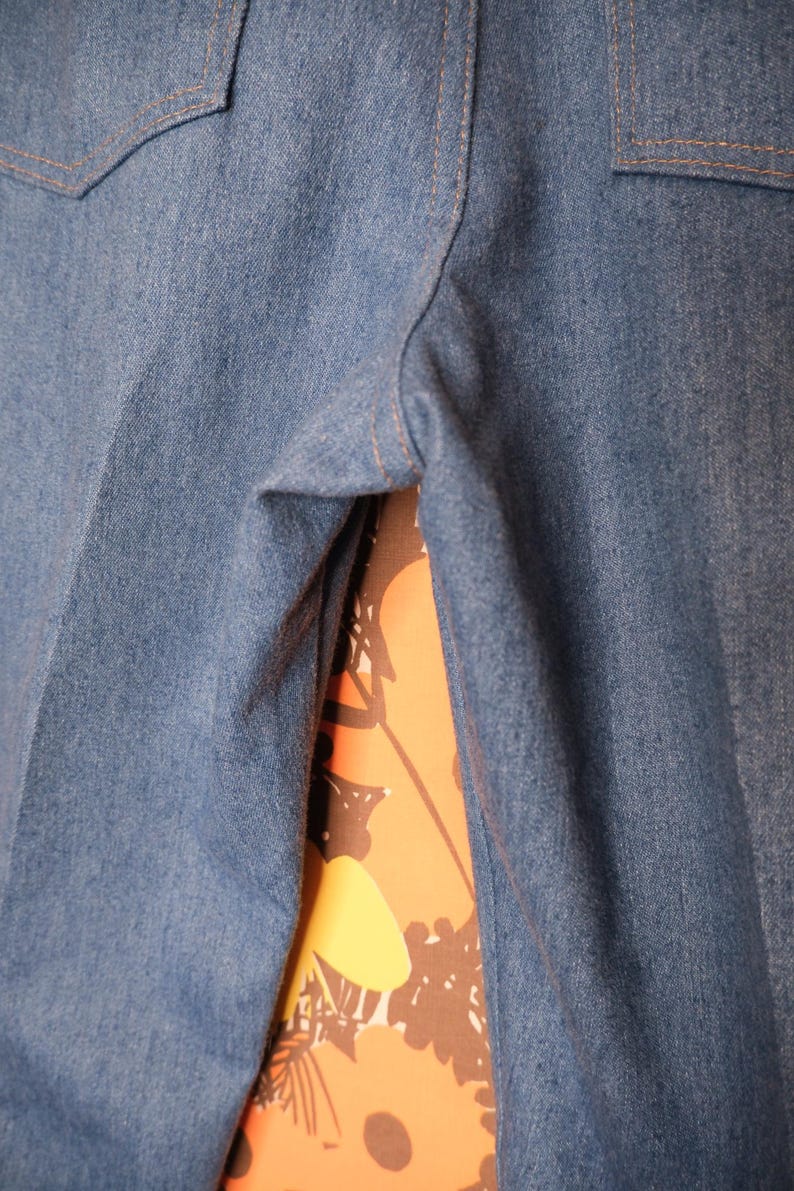May include: Close-up of blue denim pants with two pockets and tan stitching. The pants are made of a medium-weight denim fabric. The background shows a floral pattern in orange, yellow, and brown.