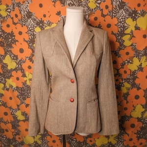 May include: A tan blazer with a herringbone pattern. The blazer has two red buttons and two front pockets. The blazer is displayed on a mannequin. The background is a floral pattern with orange, yellow, and brown flowers.