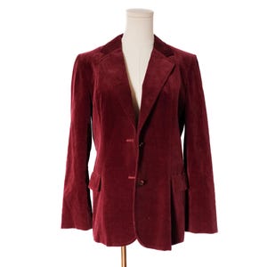 May include: A burgundy velvet blazer with a notched lapel, two-button closure, and two front pockets. The jacket is displayed on a mannequin against a white background. The blazer has long sleeves and a classic tailored fit.