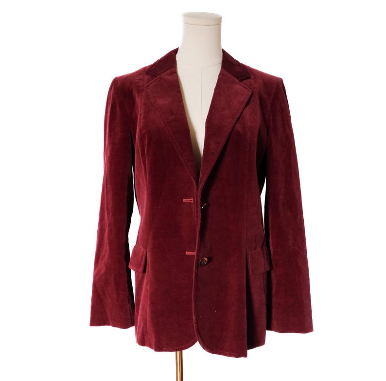 May include: A burgundy velvet blazer with a notched lapel, two-button closure, and two front pockets. The jacket is displayed on a mannequin against a white background. The blazer has long sleeves and a classic tailored fit.