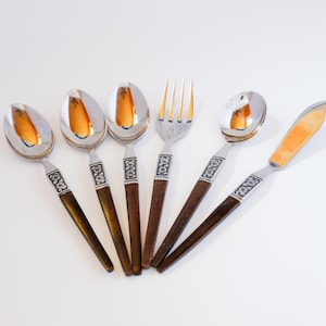 May include: A set of six vintage silverware pieces, including spoons, a fork, and a butter knife. The spoons and knife have silver blades with a gold-colored design. The handles are dark brown with silver decorative bands.