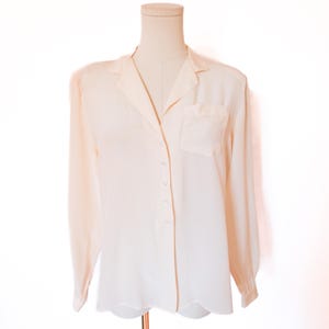 May include: A light peach-colored button-down shirt with long sleeves and a single chest pocket. The shirt is displayed on a mannequin, showcasing its classic collar and button-up design. The fabric appears to be lightweight and flowing.