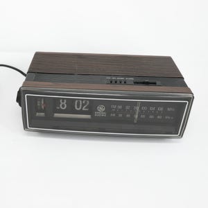 May include: A vintage General Electric digital alarm clock. The clock has a brown top with a ridged design and a black front panel displaying the time as 8:02. It features AM/FM radio frequencies and the General Electric logo.