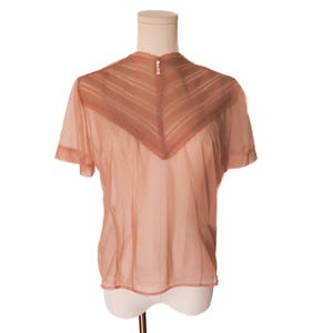 May include: A sheer, peach-colored blouse with short sleeves and a high neckline. The blouse features a decorative, V-shaped panel on the upper chest and three small buttons at the back of the neck. The garment is displayed on a mannequin.