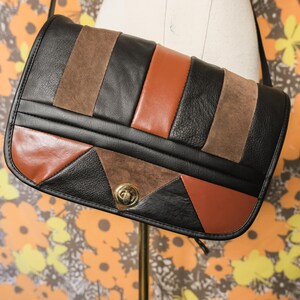 May include: A rectangular shoulder bag featuring a color-blocked design of black, brown, and tan panels. The bag is constructed with a mix of smooth and suede textures, accented by a gold-toned metal turn-lock closure on the front flap.