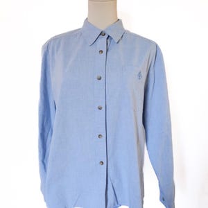 May include: A light blue button-down shirt with long sleeves, displayed on a mannequin. The shirt has a small embroidered emblem on the chest pocket and features a classic collar and buttoned cuffs. The fabric appears to be a soft, woven material.