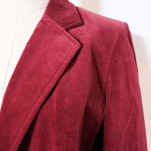 May include: A close-up of a burgundy blazer. The blazer is made of a soft, velvety material and features a classic notched lapel and a tailored fit. The color is a deep, rich shade of red.