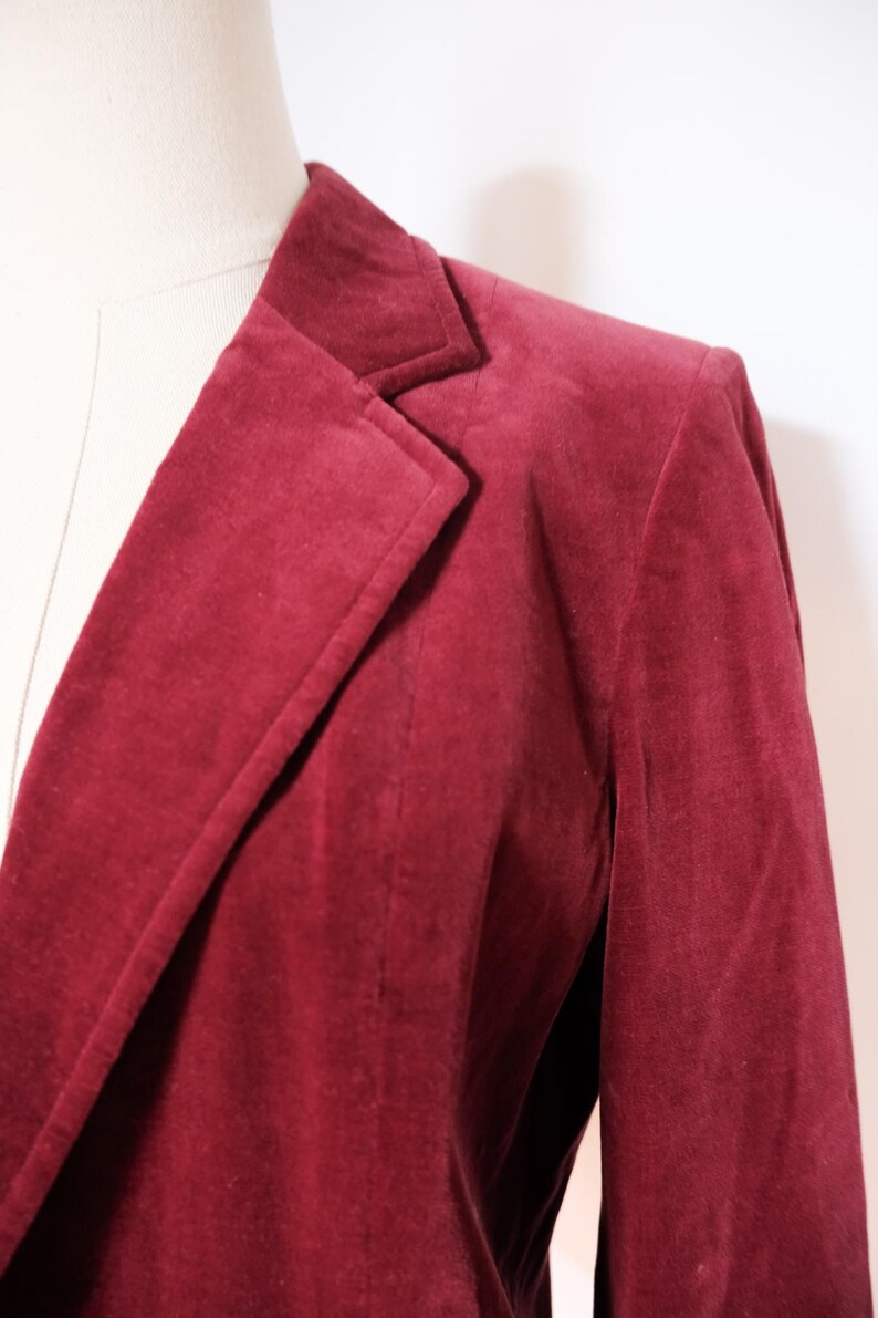 May include: A close-up of a burgundy blazer. The blazer is made of a soft, velvety material and features a classic notched lapel and a tailored fit. The color is a deep, rich shade of red.