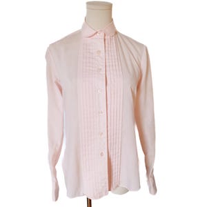 May include: Pale pink long-sleeve shirt with a pleated bib front and button-down closure. The shirt has a classic collar and cuffs. The garment is displayed on a mannequin against a white background.