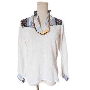 May include: White long-sleeve shirt with a textured fabric. The shirt features floral patterned accents on the shoulders, collar, and cuffs. The collar has a V-neck design with a decorative trim. The shirt is displayed on a mannequin.