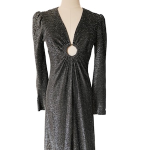 May include: A long, black, sparkly dress with long sleeves and a plunging V-neckline. The dress features a circular cutout detail at the bust and subtle shoulder pads. The fabric has a shimmering, textured appearance, creating a glamorous look.