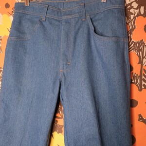 May include: Blue denim pants with a classic five-pocket design, hanging on a clear plastic hanger. The pants have a straight leg cut and are made of a medium-wash denim fabric. The stitching is a contrasting orange color.