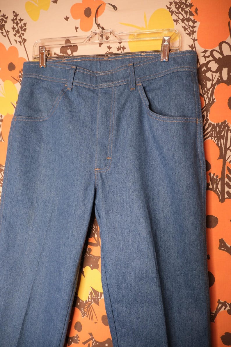 May include: Blue denim pants with a classic five-pocket design, hanging on a clear plastic hanger. The pants have a straight leg cut and are made of a medium-wash denim fabric. The stitching is a contrasting orange color.