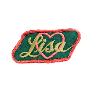 May include: Embroidered patch with the name "Lisa" in gold script, set within a pink heart on a dark green background. The patch has a red, zig-zag border, and is rectangular with rounded corners.