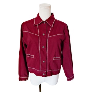 May include: A deep burgundy jacket featuring a classic collar, long sleeves, and a front button closure. The garment is accented with prominent white zigzag stitching along the seams, pockets, and cuffs, providing a distinct contrast.