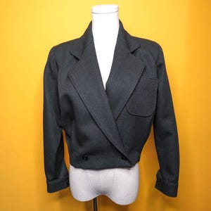 May include: A dark gray cropped blazer with a double-breasted design. The jacket features a notched lapel, a single pocket, and two black buttons. The sleeves are cuffed, and the blazer is displayed on a mannequin against a yellow background.