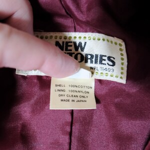 May include: Close-up of a tag from a garment. The tag reads "NEW STORIES" with "WPL 11403" below. The tag also states "SHELL: 100% COTTON, LINING: 100% NYLON, DRY CLEAN ONLY, MADE IN JAPAN". The tag is held over a burgundy fabric.
