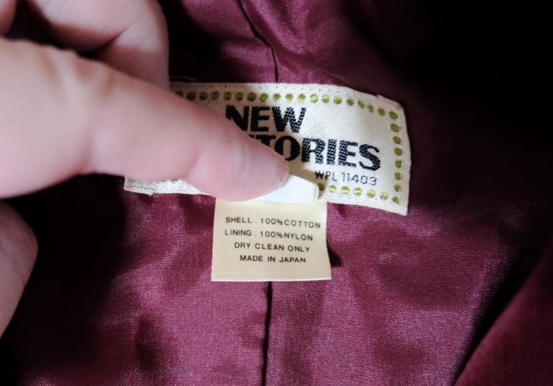 May include: Close-up of a tag from a garment. The tag reads "NEW STORIES" with "WPL 11403" below. The tag also states "SHELL: 100% COTTON, LINING: 100% NYLON, DRY CLEAN ONLY, MADE IN JAPAN". The tag is held over a burgundy fabric.