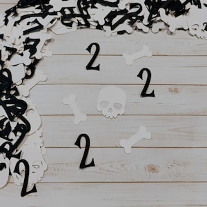 May include: A Halloween-themed arrangement featuring black and white cutouts of skulls, bones, and the number "2". The cutouts are scattered on a light-colored wooden surface, creating a festive and spooky atmosphere.