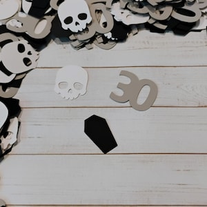 May include: A collection of Halloween-themed paper cutouts, including skulls, coffins, and the number "30". The cutouts are in shades of black, white, and gray, scattered on a light wood-grain surface. The image has a festive, spooky aesthetic.