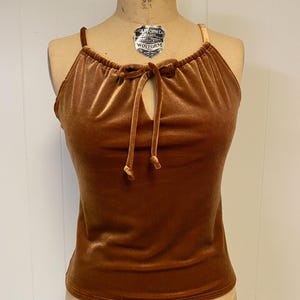 May include: A sleeveless top crafted from brown velvet fabric. The garment features a gathered neckline with a central tie closure that creates a small keyhole opening. The top has thin shoulder straps and a straight hemline at the waist.