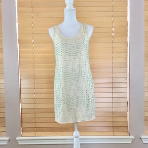 May include: A sleeveless, cream-coloured dress with a beaded design. The dress is on a white dress form and the dress form is on a white stand.