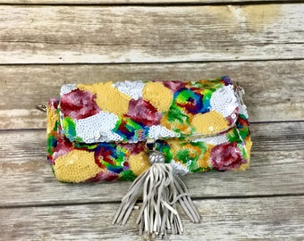 Può includere: Una piccola pochette di paillettes colorata con un fiocco bianco. La borsa è decorata con una varietà di colori, tra cui rosso, giallo, verde, blu e viola.