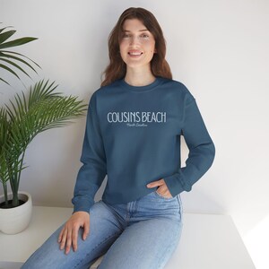 Peut inclure: Un sweat-shirt bleu sarcelle avec le texte "COUSINS BEACH North Carolina" en blanc. Le haut à col ras du cou et à manches longues est porté par une personne en jean bleu. Une plante verte dans un pot blanc est en arrière-plan.