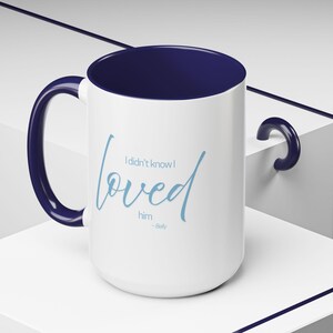 May include: White ceramic mug with a navy blue interior, handle, and rim. The mug features the light blue text "I didn't know I loved him - Belly" in a cursive font. The mug is on a white surface.