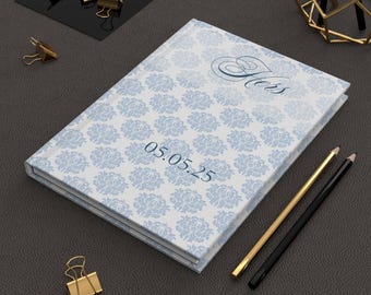 May include: A hardcover journal with a light blue floral pattern and the word "Hers" in script. The date "05.05.25" is printed on the cover. Gold and black pencils and gold binder clips are also visible.