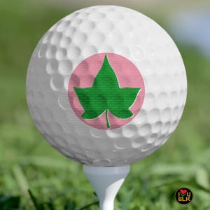 May include: A white golf ball with a pink circle and a green ivy leaf design, resting on a white tee. The golf ball is on a green grassy surface.