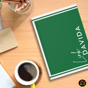 May include: A green notebook with white border lines and the name "Linda Davida" in white script. The notebook is on a wooden desk with a cup of coffee, a keyboard, and a notepad.