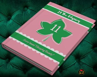 May include: A pink notebook with a green ivy leaf design and the number 11 in the center. The text "21 The Odyssey" and "Soror Imani "Sleeping Beauty"" is printed on the cover.
